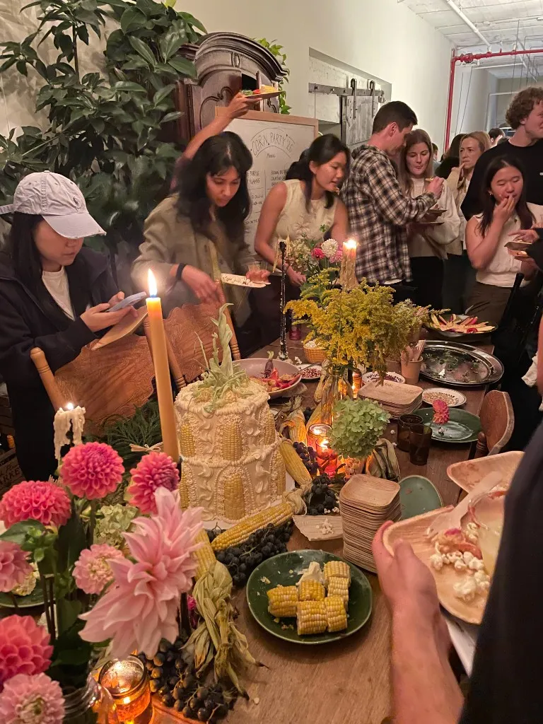 A candlelit table set with a corn cake, dahlias, and plates of corn, with guests gathered behind it.