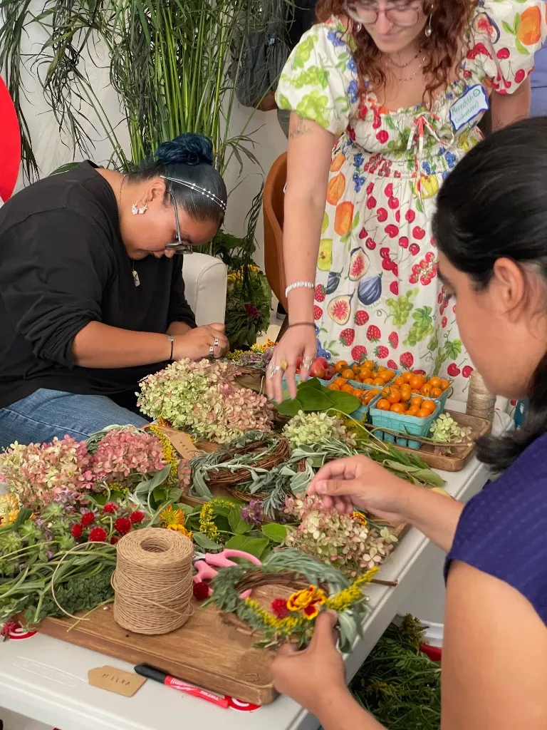 Three people at a table assembling small wreaths from hydrangea, sage, and other botanicals, with twine, scissors, and punnets of fruit.