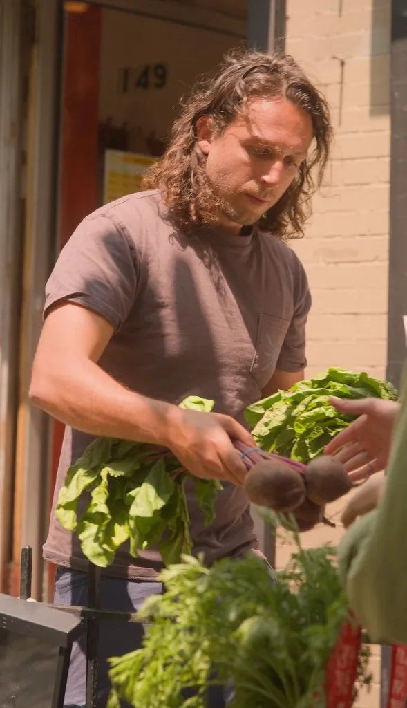 A grower handing a bunch of beets and greens to a customer at a sidewalk farmstand.