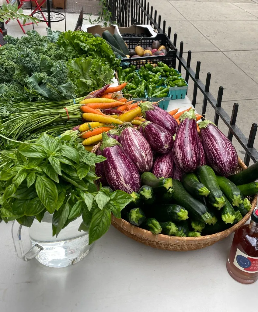 A farmstand table crowded with eggplants, zucchini, carrots, basil, and a black crate of blackberries, set up along a sidewalk railing.