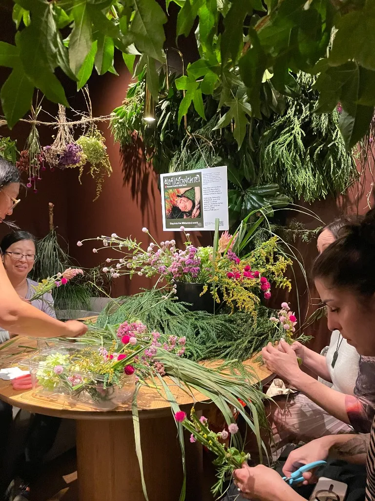 Guests arranging wildflowers and herbs into bouquets at a round table under hanging greenery.
